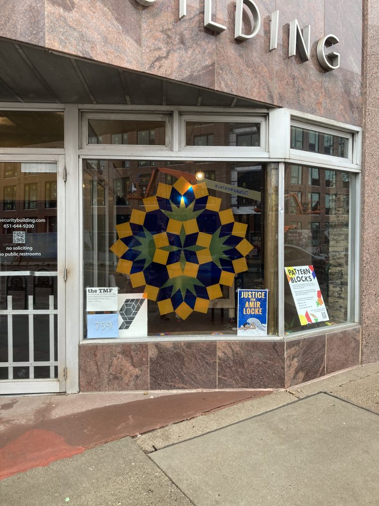 Large window with a large symmetrical arrangement of squares and diamonds in yellow, blue and orange. The street-level window is viewed from the sidewalk