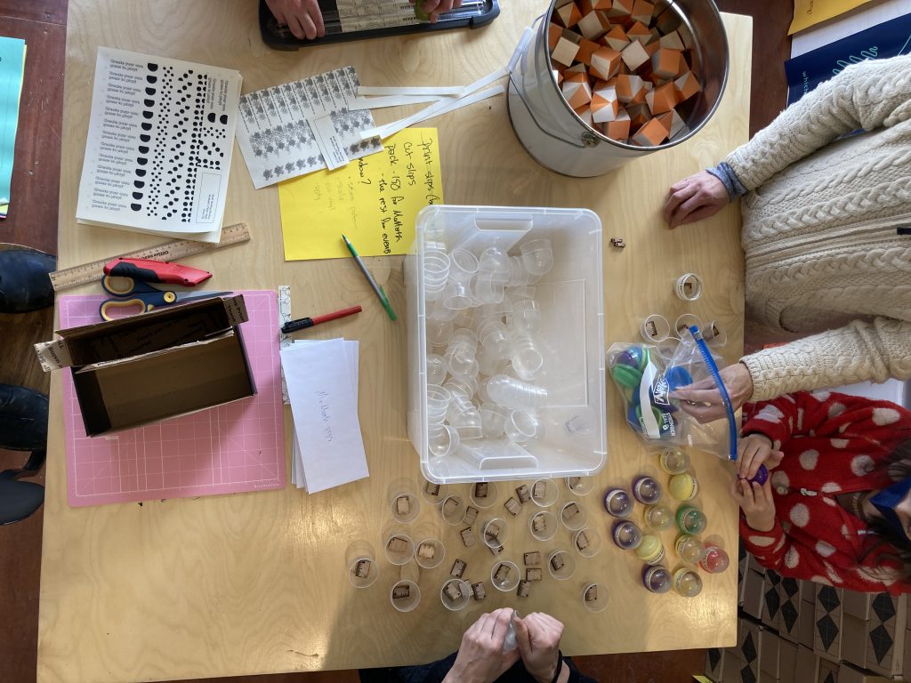 Overhead view of a large square wooden table. There are colorful blocks, scissors, paper, and a collection of vending machine capsules. Four people stand at the table organizing the materials.