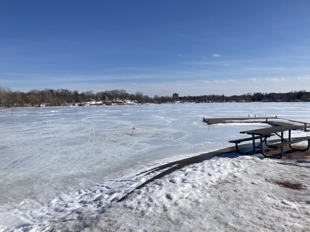 A view across a frozen lake. The sky is blue with thin clouds on the horizon. There are tracks—perhaps snowmobile–coming fron shore and curving gracefully in many directions across the ice. 