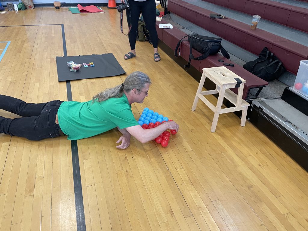 I am lying on my belly on the wooden floor of a gymnasium, carefully arranging brightly colored blue and red plastic balls into a diamond shape. In the background stands a photographer in sandals and an arrangement of dice on black butcher paper.