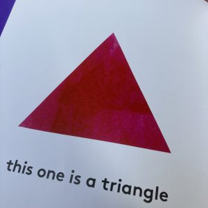 A page from a shapes book. There is a beautiful, bold, red, equilateral triangle in the center of the page, with a horizontal base. The words underneath read, "this one is a triangle"