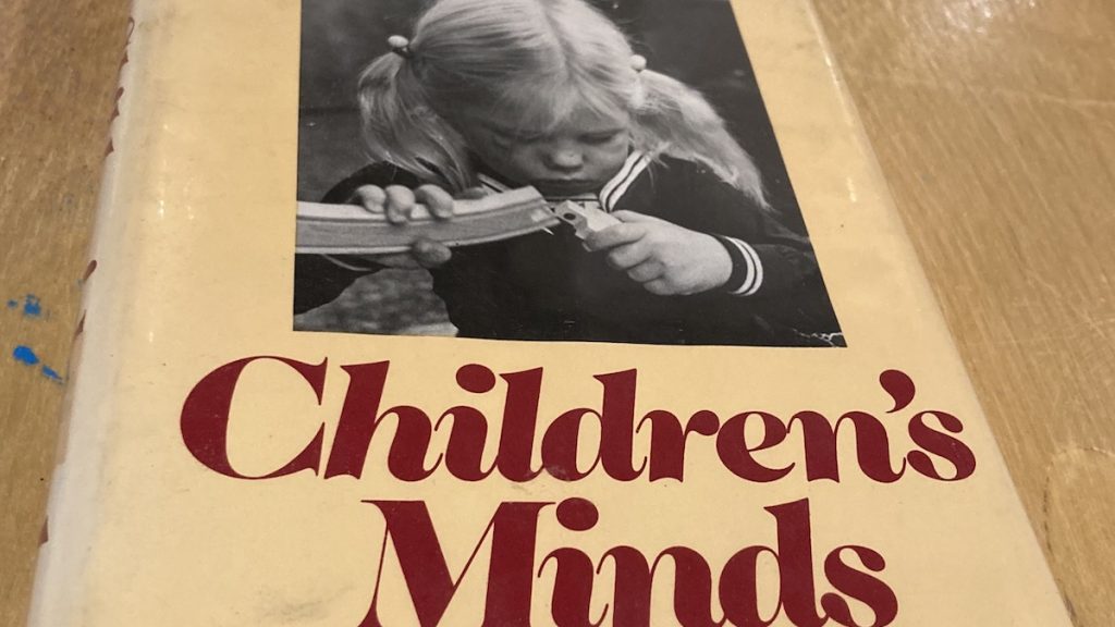 The cover of the book "Children's Minds". It contains a photo of a young girl intently studying two pieces of wooden track for a toy train, as she attempts to assemble them. This is a beautiful illustration of the relationship between children's minds and their physical environments.