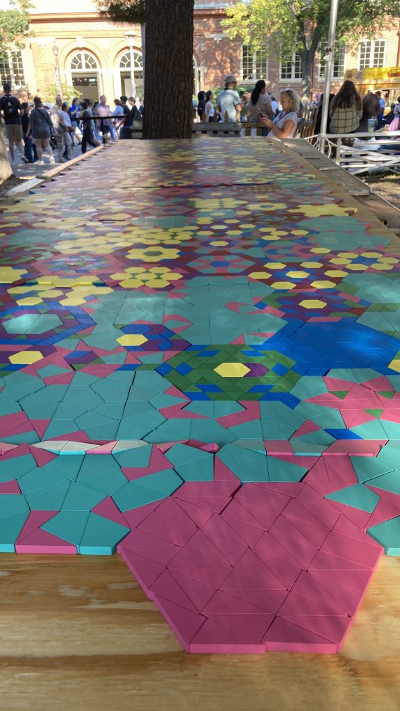 A deep-field shot of the mosaic. There is a large hexagon in the foreground, composed entirely of pink right triangles. Hexagons—tiled together—recede into the distance; each composed of small, brightly colored wooden polygons.