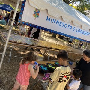 Children stand at a table assembling hexagons from pattern blocks. A tent with banner reading "Minnesota's Largest [obscured]" is in the background.