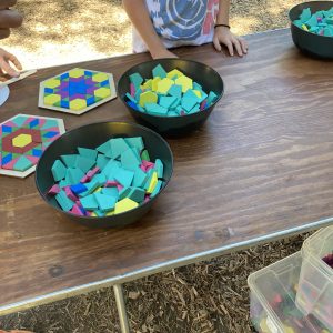 A wood folding table holds several black plastic bowls full of colorful wooden polygons. There are two large hexagons composed of these smaller shapes on the table, and a young person standing nearby.