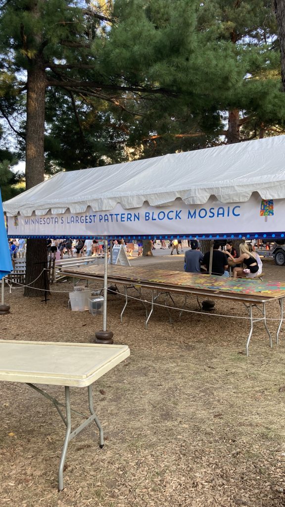 A long, thing, white event tent with a banner reading "Minnesota's Largest Pattern Block Mosaic". Inside the tent are folding tables, upon which rest wooden boards and a mosaic of many many pattern blocks.